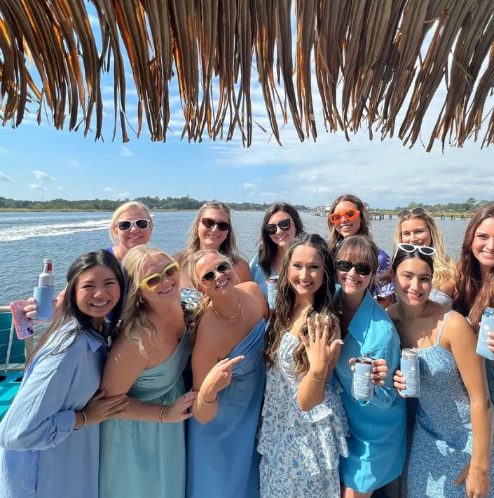 Smiling group of friends in coordinating blue outfits and sunglasses posing on a boat under a thatched canopy, holding drinks with a sunny waterfront bay and pier in the background.