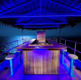 Tiki-style bar on a boat deck at night — bamboo counter, round stools and blue LED lights under a thatched roof overlooking the moonlit ocean.