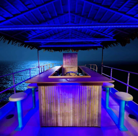 Tiki-style bar on a boat deck at night — bamboo counter, round stools and blue LED lights under a thatched roof overlooking the moonlit ocean.