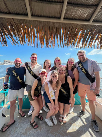 Cheerful bachelorette crew posing on a boat under a thatched tiki roof, bride in a white sash and veil with friends wearing black sashes, sunny blue-sky marina and calm water in the background.