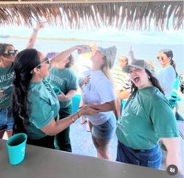 Friends celebrating on a sunny Charleston-area boat cruise, women in matching green tees and summer hats laughing under a thatched canopy as one takes a drink with water and shoreline in the background.