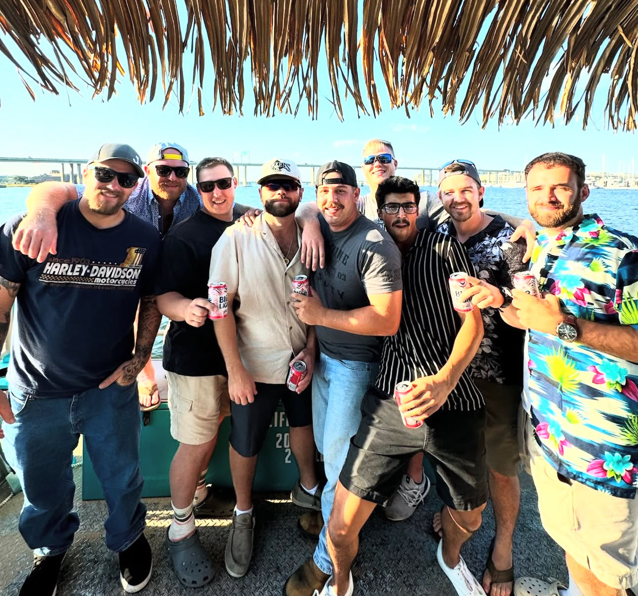 Group of nine men in casual summer clothes and sunglasses on a boat deck under a thatched tiki roof, holding beer cans with a bridge and blue water in the sunny background — lively waterfront boat party scene.