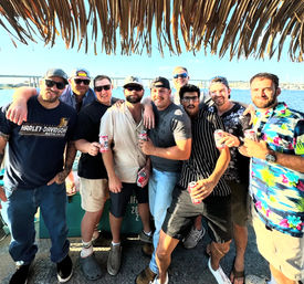 Group of nine men in casual summer clothes and sunglasses on a boat deck under a thatched tiki roof, holding beer cans with a bridge and blue water in the sunny background — lively waterfront boat party scene.