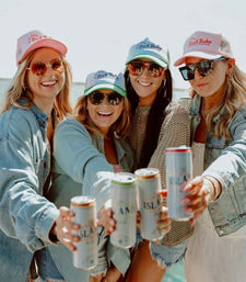 Four friends in trucker hats and sunglasses toasting canned drinks on a sunny coastal boat, summer beach vibe.