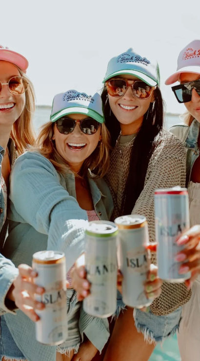 Four friends in trucker hats and sunglasses toasting canned drinks on a sunny coastal boat, summer beach vibe.