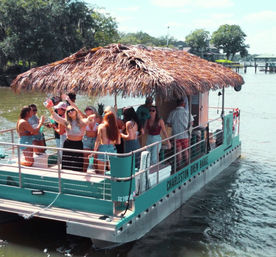 Group of people partying on a teal pontoon boat with a thatched tiki roof cruising a sunny river past tree-lined shore and docks