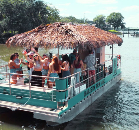 Group of people partying on a teal pontoon boat with a thatched tiki roof cruising a sunny river past tree-lined shore and docks