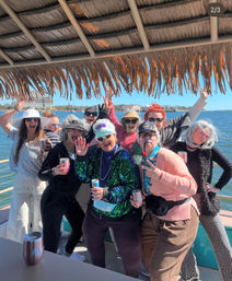 Cheerful group in playful costumes and sunglasses posing under a thatched tiki roof on a sunny boat cruise along a coastal waterfront with city buildings across the bay, holding drinks and cheering.