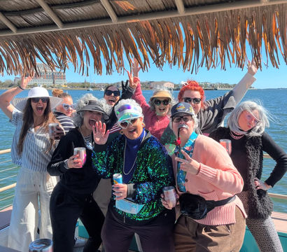 Cheerful group in playful costumes and sunglasses posing under a thatched tiki roof on a sunny boat cruise along a coastal waterfront with city buildings across the bay, holding drinks and cheering.
