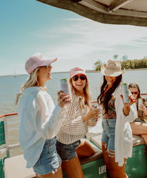 Three friends in summer hats and denim shorts laughing and toasting canned drinks on a sunny boat cruise on a lake with trees and sailboats in the distance