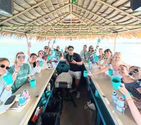 Group of friends raising turquoise cups aboard a tiki-roof pontoon boat on calm coastal waters, daytime celebration and smiling faces.