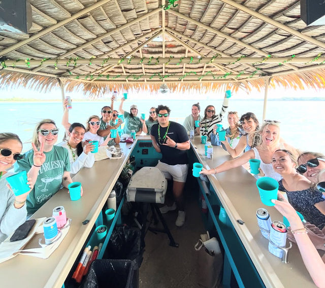 Group of friends raising turquoise cups aboard a tiki-roof pontoon boat on calm coastal waters, daytime celebration and smiling faces.