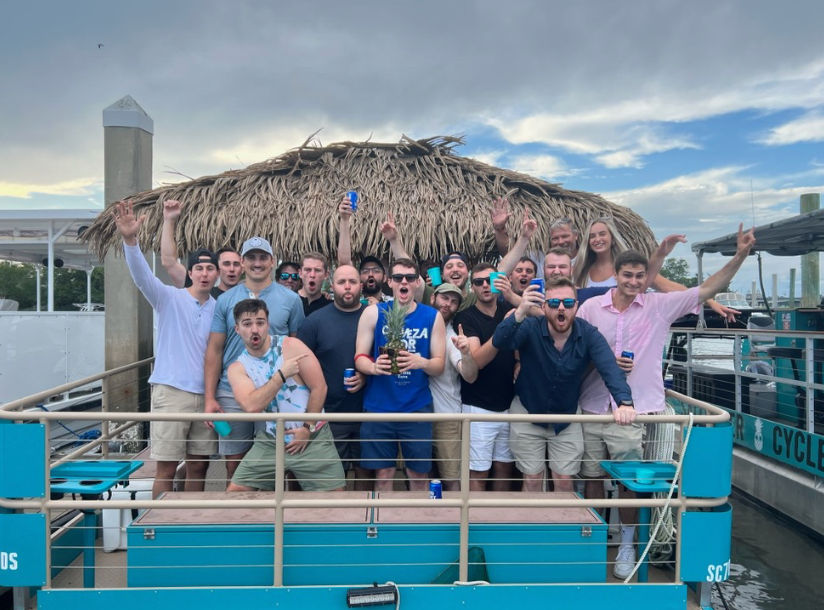 Group of friends cheering with drinks on a turquoise boat deck beneath a thatched tiki hut at a marina, cloudy sky and docked boats in the background