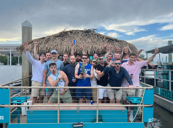 Group of friends cheering with drinks on a turquoise boat deck beneath a thatched tiki hut at a marina, cloudy sky and docked boats in the background