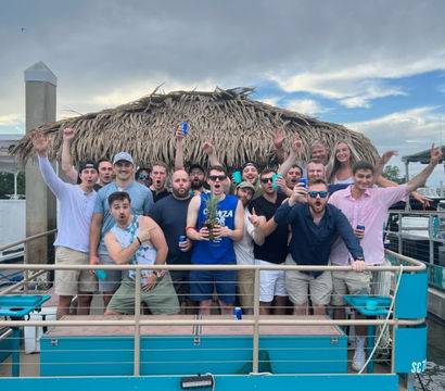 Group of friends cheering with drinks on a turquoise boat deck beneath a thatched tiki hut at a marina, cloudy sky and docked boats in the background