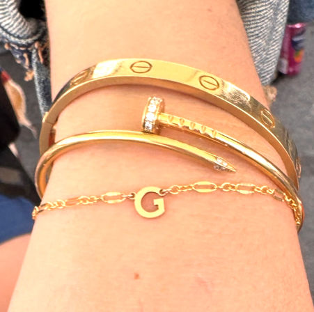 Close-up of a wrist with stacked gold bracelets: hammered bangle, rhinestone-accented nail-shaped cuff, smooth bangle, and delicate chain with letter G charm.