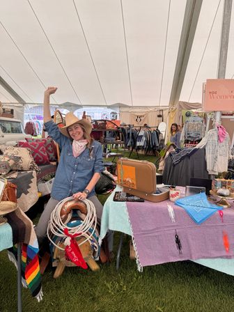 Smiling woman in a cowboy hat and denim shirt waves from a country-fair market tent booth selling western gear — saddle and lariat, colorful blankets and bandanas, vintage suitcase and clothing racks on grass.