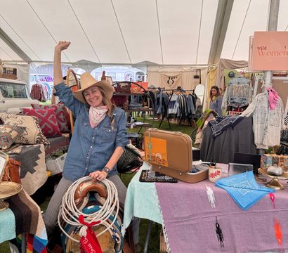 Smiling woman in a cowboy hat and denim shirt waves from a country-fair market tent booth selling western gear — saddle and lariat, colorful blankets and bandanas, vintage suitcase and clothing racks on grass.