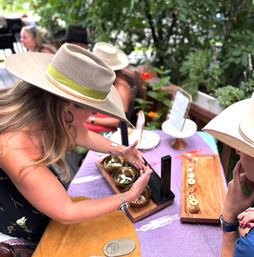 Person in a wide-brim hat demonstrating brass singing bowls and handmade jewelry at an outdoor artisan market table with a purple cloth, wooden trays and potted plants