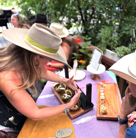 Person in a wide-brim hat demonstrating brass singing bowls and handmade jewelry at an outdoor artisan market table with a purple cloth, wooden trays and potted plants