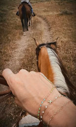 View from horseback: hand wearing delicate beaded bracelets holding the reins of a chestnut horse with a white mane, following another rider down a dry grassy country trail — outdoor trail ride.