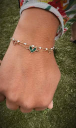 Close-up of a person’s wrist wearing layered delicate gold bracelets with a green heart charm and tiny white beads, floral-print sleeve and grassy outdoor background.