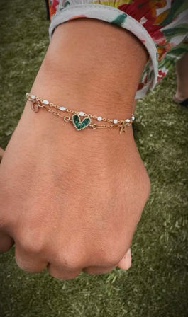 Close-up of a person’s wrist wearing layered delicate gold bracelets with a green heart charm and tiny white beads, floral-print sleeve and grassy outdoor background.