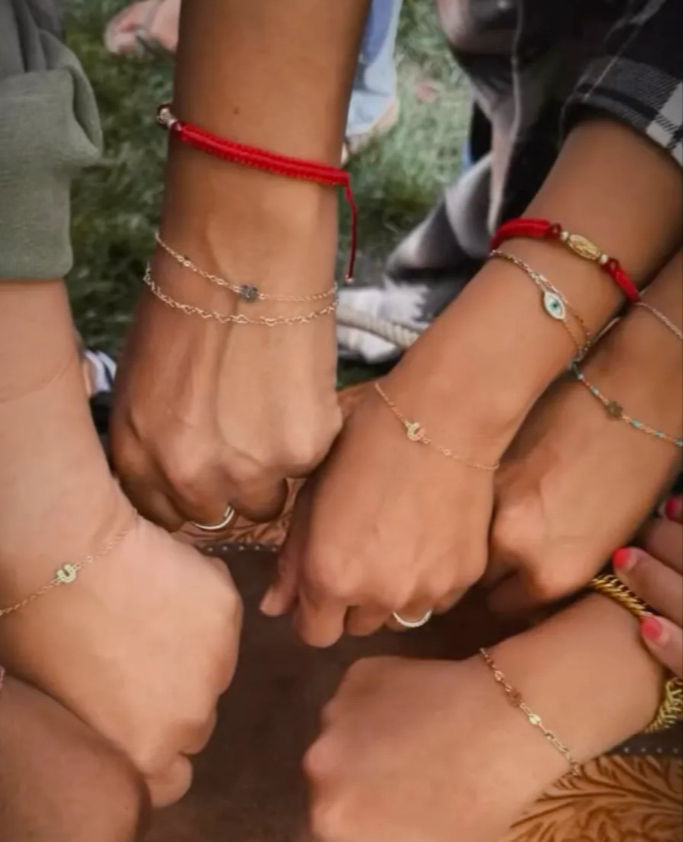 Close-up of six friends' joined fists in a circle outdoors, showcasing red-string and delicate gold friendship bracelets with evil-eye and charm pendants.