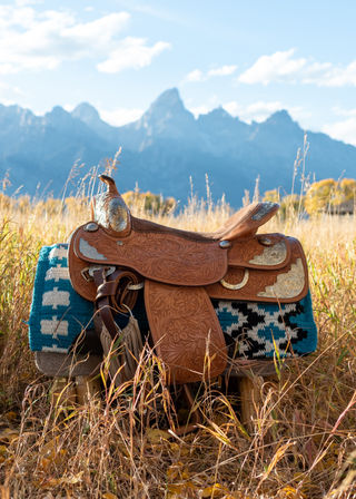 Tooled leather western saddle on a turquoise patterned saddle blanket in a golden autumn meadow with jagged blue mountain peaks in the background