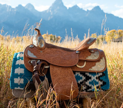 Tooled leather western saddle on a turquoise patterned saddle blanket in a golden autumn meadow with jagged blue mountain peaks in the background