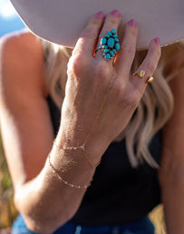 Sunlit outdoor close-up of a hand holding a beige wide-brim hat, featuring a turquoise cluster ring, gold rings and delicate hand-chain bracelets — boho summer jewelry style.