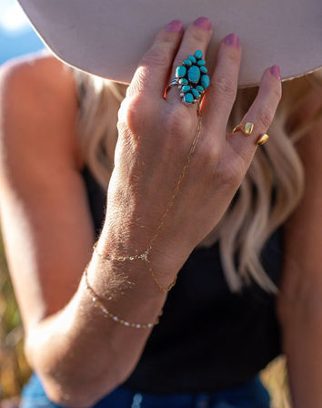 Sunlit outdoor close-up of a hand holding a beige wide-brim hat, featuring a turquoise cluster ring, gold rings and delicate hand-chain bracelets — boho summer jewelry style.