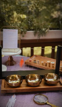 Three hammered brass bowls on a wooden tray on a sunlit garden deck table, with a magnifying glass and craft display board, green plants in the background