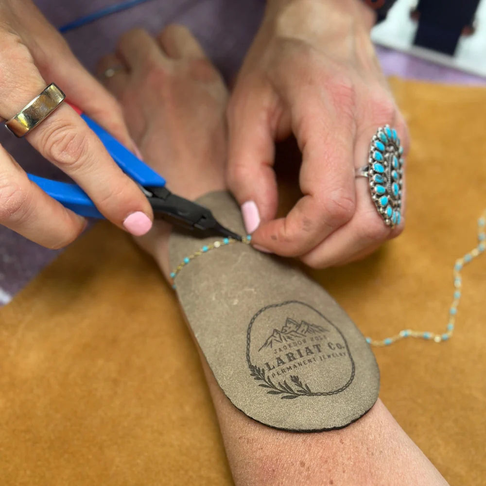 Hands using blue-handled pliers to fasten a delicate turquoise beaded bracelet on a wrist over a leather pad; turquoise cluster ring and pink nails visible in an artisan jewelry studio.