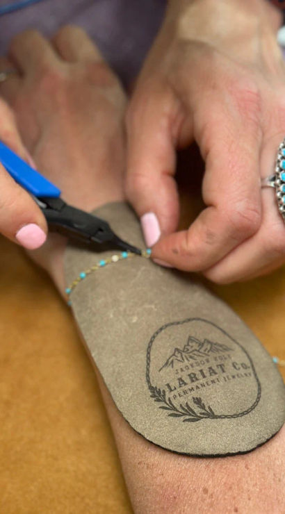Hands using blue-handled pliers to fasten a delicate turquoise beaded bracelet on a wrist over a leather pad; turquoise cluster ring and pink nails visible in an artisan jewelry studio.
