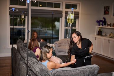 At-home IV therapy in a cozy living room: a clinician in black scrubs adjusts an IV drip for a woman reclining on a gray sofa, with IV bags on poles and friends chatting in the background.