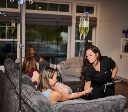 At-home IV therapy in a cozy living room: a clinician in black scrubs adjusts an IV drip for a woman reclining on a gray sofa, with IV bags on poles and friends chatting in the background.