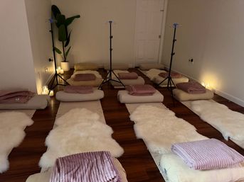 Candlelit meditation setup with rows of floor cushions and bolsters, cream sheepskin rugs and mauve throw blankets on a hardwood floor, a potted plant and three black tripod stands — cozy relaxation/wellness space.