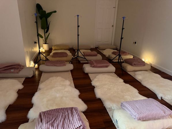 Candlelit meditation setup with rows of floor cushions and bolsters, cream sheepskin rugs and mauve throw blankets on a hardwood floor, a potted plant and three black tripod stands — cozy relaxation/wellness space.