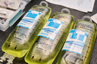 Three translucent IV saline bags with blue labels and measurement markings, yellow-tinted solution laid on a clinic countertop next to a medical supply kit — ready for infusion.