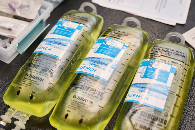 Three translucent IV saline bags with blue labels and measurement markings, yellow-tinted solution laid on a clinic countertop next to a medical supply kit — ready for infusion.