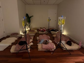 Dim, candlelit wellness studio with people reclining on floor cushions and blankets while IV infusion bags on stands hold yellow fluid; cozy pillows, a potted plant, and soft lighting create a relaxed, spa-like treatment room.
