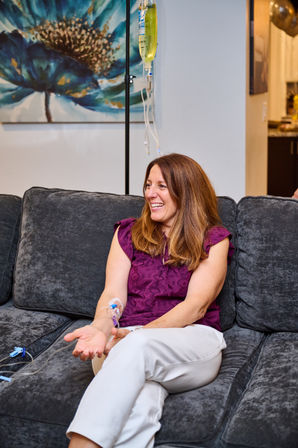 Woman smiling on gray sofa receiving at-home IV hydration therapy — yellow IV bag on stand connected to a catheter in her forearm, decorative blue floral painting in the background.