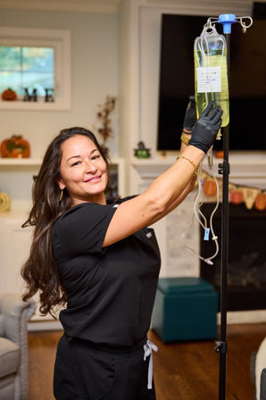 Healthcare worker in black scrubs and gloves adjusts an IV hydration bag on a stand in a cozy living room with autumn decorations.