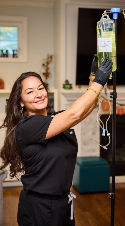 Healthcare worker in black scrubs and gloves adjusts an IV hydration bag on a stand in a cozy living room with autumn decorations.