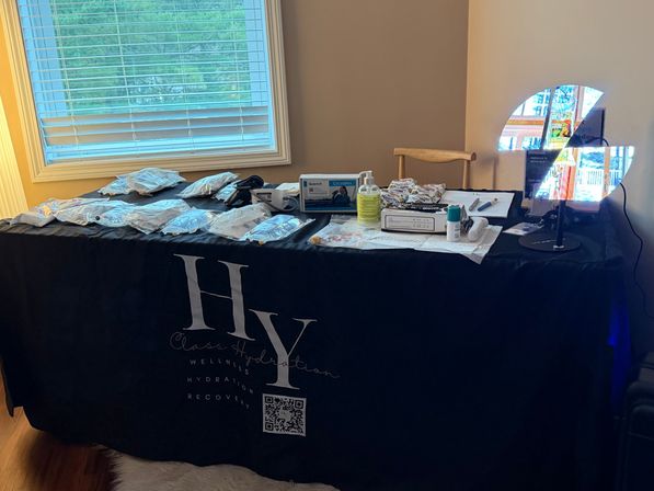 Indoor wellness hydration station by a window: black-covered table displaying IV bags, tubing, sanitizer, boxes, snacks and a small LED display.