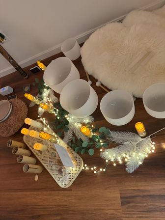 Cozy indoor sound bath setup on hardwood floor with white crystal singing bowls, mallets, LED candles, fairy lights, eucalyptus, crystals and a fluffy sheepskin rug for meditation