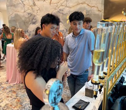 Young adults sampling drinks at a gold-framed beverage tasting station inside a stylish hotel lobby event with a veined marble wall backdrop and patterned carpet