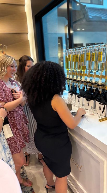 Women sampling fragrances at a boutique perfume bar — row of glass dispensers pouring golden perfume into sample bottles beside gift bags.