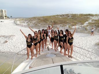 Cheerful bachelorette group in matching "Bride Squad" swimsuits raising champagne on the bow of a boat at a white‑sand beach with grassy dunes and calm ocean in the background.